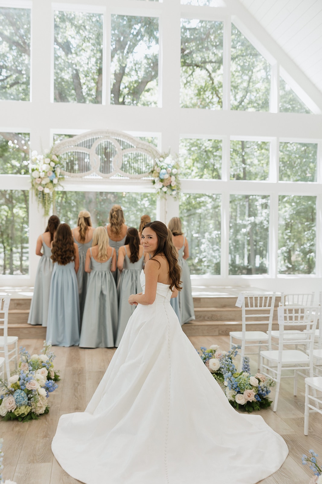 Nina in her strapless white ballgown glancing back over her shoulder as her bridesmaids in French blue dresses face the antique arch inside The French Farmhouse chapel