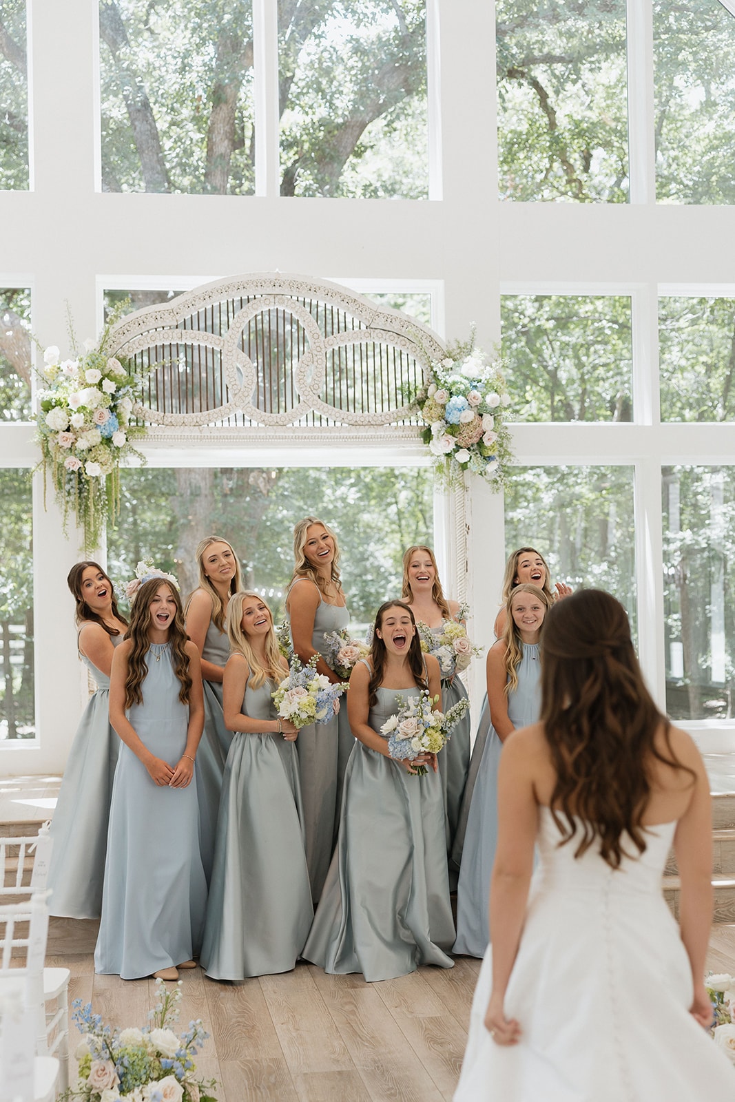 Nina's bridesmaids in French blue dresses reacting with pure joy and laughter as they see her in her wedding gown for the first time inside The French Farmhouse chapel