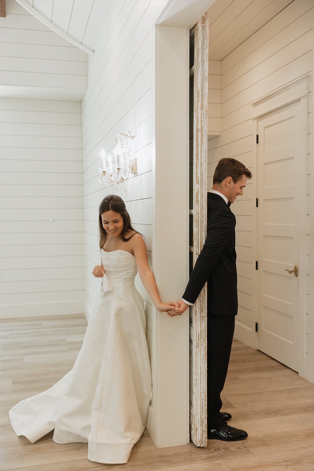Nina and Gage sharing a private first touch before their ceremony at The French Farmhouse Venue — holding hands around a doorframe, both smiling, crystal chandelier and shiplap wall behind them