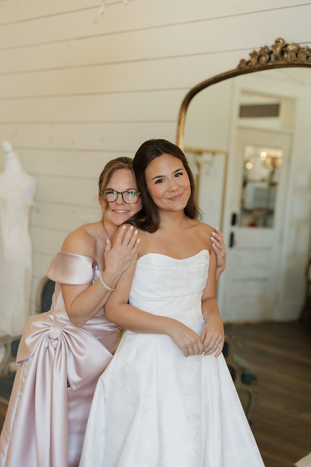 Nina and her mom Amy smiling together in the bridal suite at The French Farmhouse Venue — Amy in her blush off-the-shoulder gown with a bow, Nina in her strapless white ballgown
