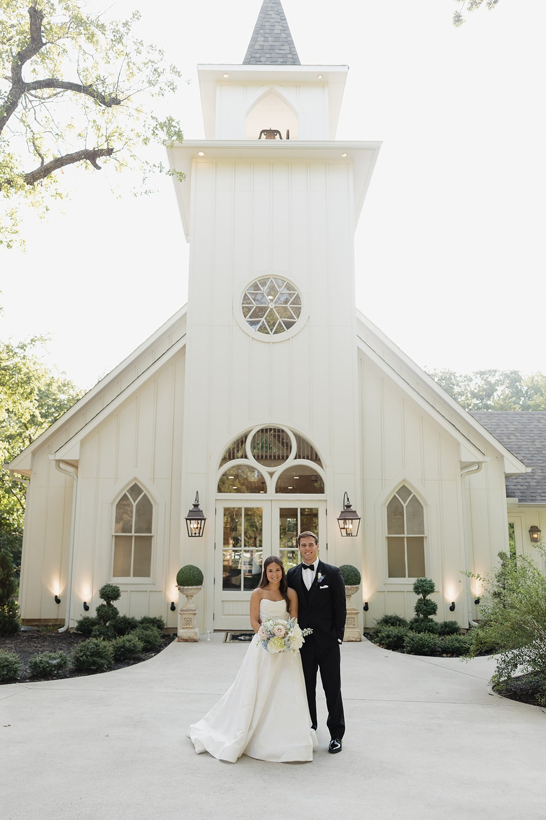 Nina and Gage standing together in front of The French Farmhouse chapel exterior — Nina in her strapless ballgown with bouquet, Gage in a black tuxedo, the white steeple and arched doors glowing behind them