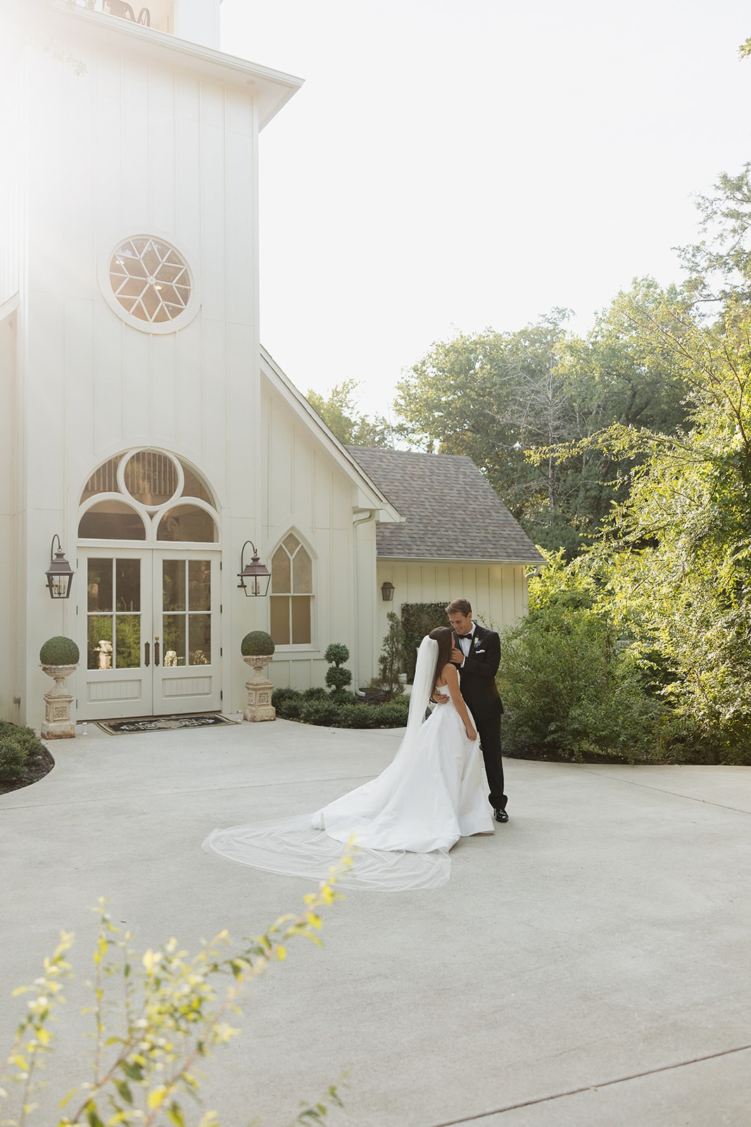 Nina and Gage sharing a romantic embrace in front of The French Farmhouse chapel — Nina's cathedral veil pooling across the courtyard, golden afternoon light flooding in from the side, summer trees surrounding them