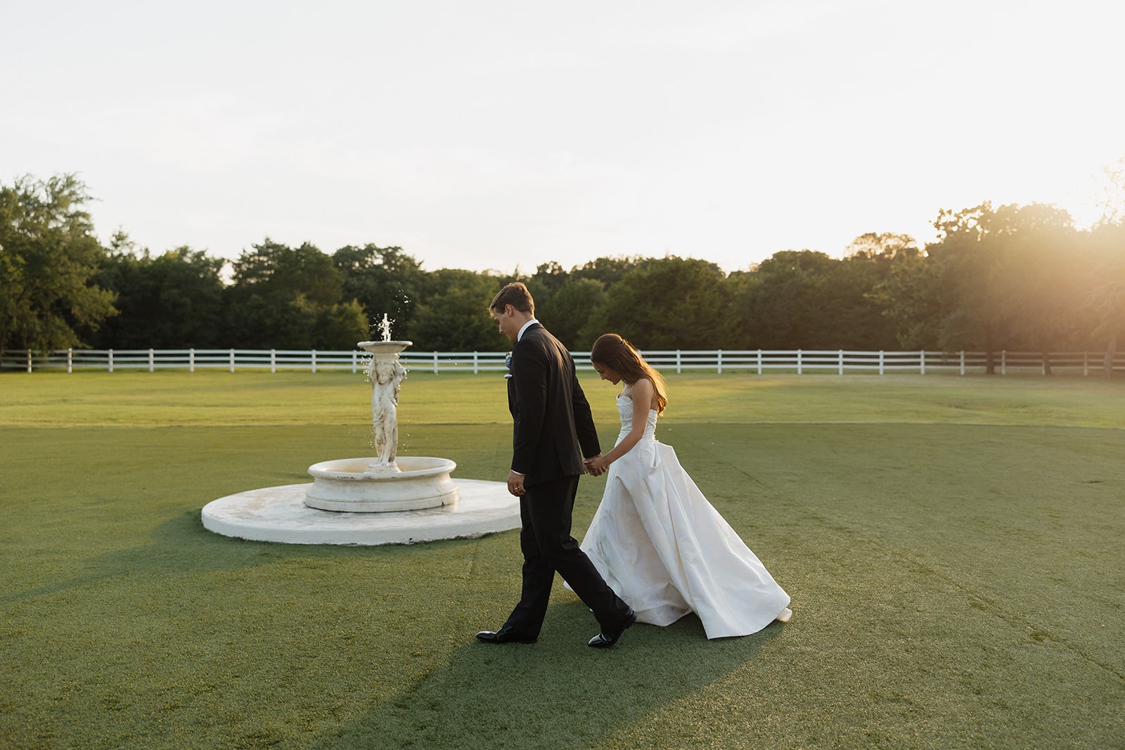 Nina and Gage walking hand in hand past the fountain on the putting green at The French Farmhouse Venue during golden hour portraits — Nina's ballgown trailing across the grass, warm Texas sunset light flooding in from behind the treeline