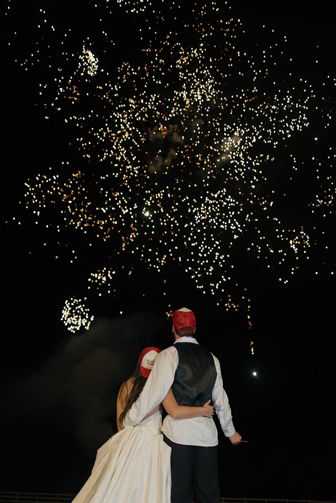 Nina and Gage in their 'Forever Ever The Jacksons' trucker hats watching a massive golden bokeh fireworks explosion fill the entire sky above The French Farmhouse Venue on their 4th of July wedding night