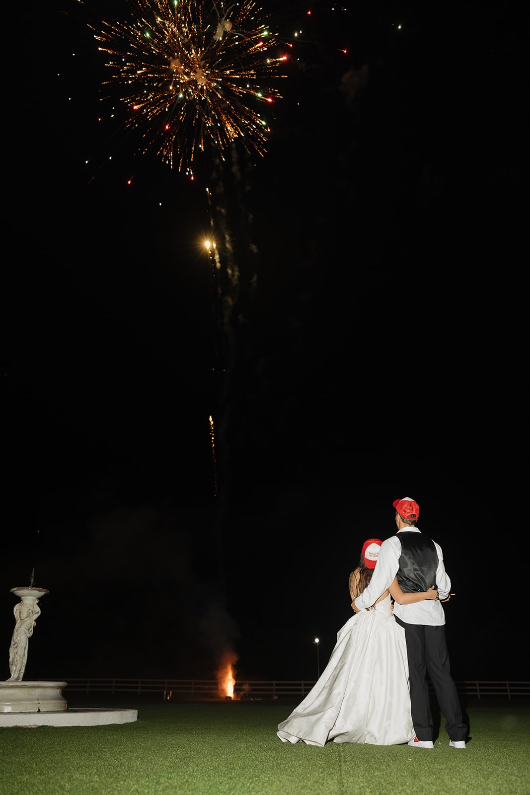 Nina and Gage standing arm in arm at the fountain on the putting green watching Dallas Elevate fireworks explode in the sky above The French Farmhouse Venue on their 4th of July wedding night