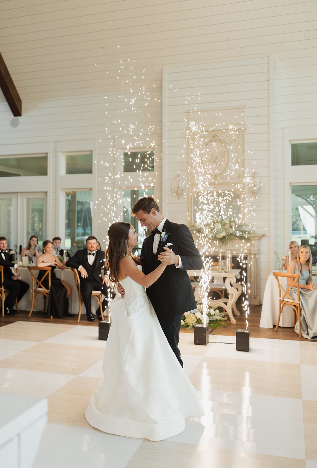 Nina and Gage sharing their first dance on the Inkredible checkerboard dance floor at The French Farmhouse Venue — Dallas Elevate cold sparks exploding behind them, the ornate fireplace mantel and shiplap walls in the background, every guest watching
