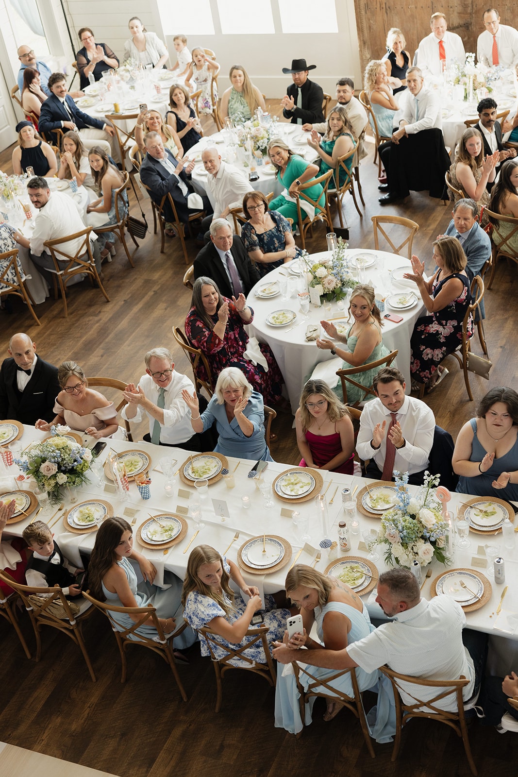 Aerial view of Nina and Gage's reception hall full of guests cheering and waving at The French Farmhouse Venue — long farm tables and round tables with blue and white floral china, JG Floral centerpieces, and the checkerboard dance floor visible in the corner