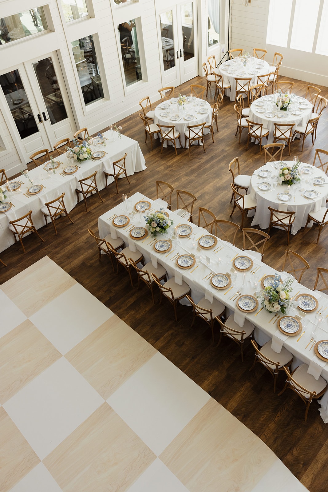 Aerial view of the reception hall at The French Farmhouse Venue set up for Nina and Gage's wedding — long farm tables and round tables with white linens, rattan chargers, blue and white floral china, gold flatware, JG Floral centerpieces, and the Inkredible checkerboard dance floor