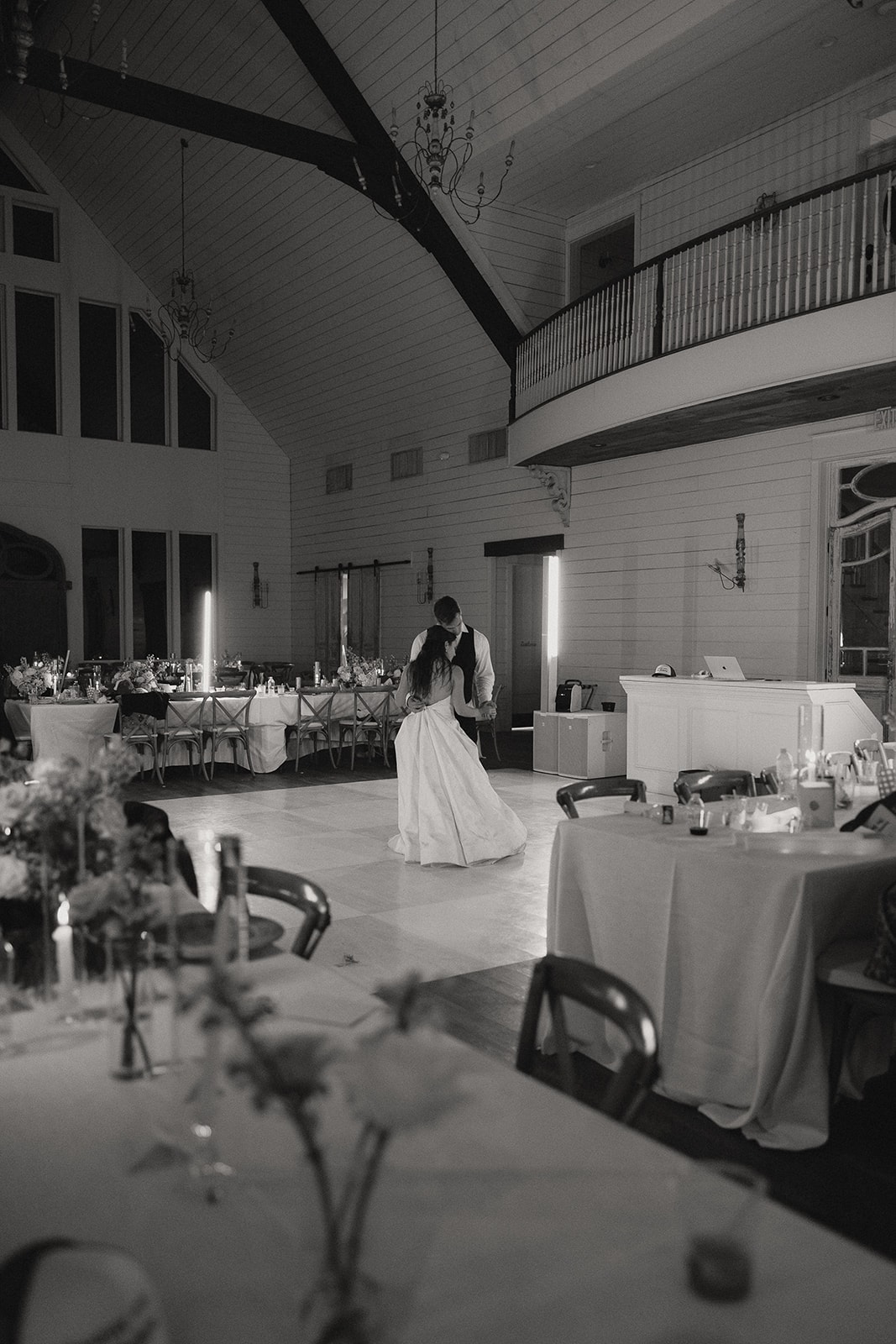 Black and white photo of Nina and Gage's private last dance alone in the empty reception hall at The French Farmhouse Venue — the checkerboard dance floor, long farm tables with candles, and the stunning balcony and cathedral ceilings above them
