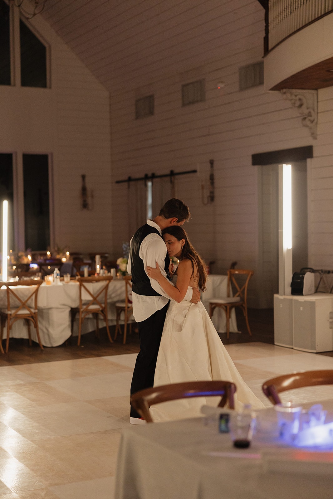 Nina and Gage sharing a private last dance alone on the Inkredible dance floor at The French Farmhouse Venue after the fireworks — candles glowing on the tables, soaring shiplap ceilings and balcony above them