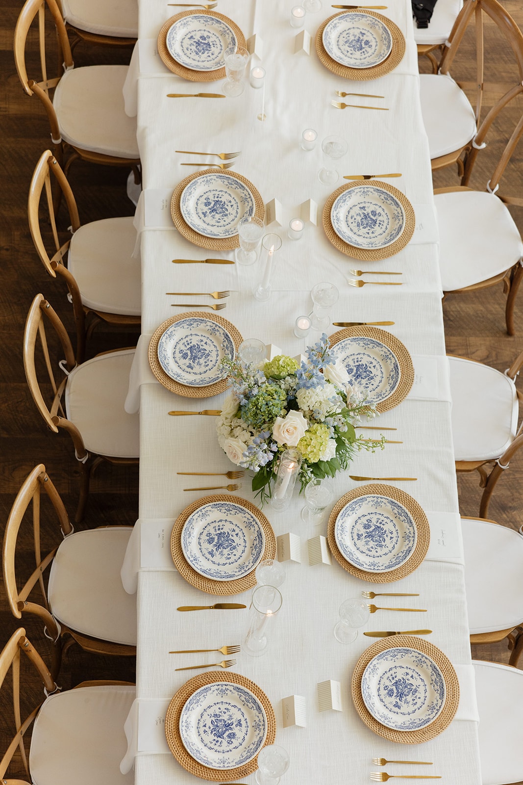 Close-up overhead detail of the reception tablescape at Nina and Gage's wedding — blue and white floral china on rattan chargers, gold flatware, taper candles, and a JG Floral centerpiece of blue hydrangeas, white roses, and green blooms