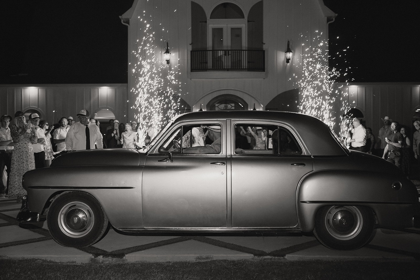 Black and white photo of Nina and Gage driving away in Lucy the antique car at The French Farmhouse Venue — guests lining the drive, cold sparks exploding behind the venue facade, the balcony and arched windows glowing in the background