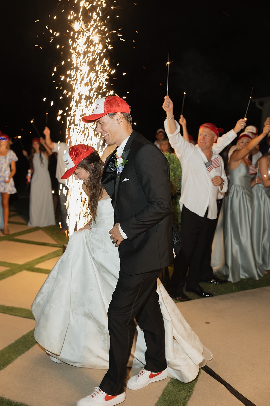 Nina and Gage walking through a tunnel of sparklers and mini American flags held by their guests at The French Farmhouse Venue send-off — both in their matching 'Forever Ever The Jacksons' trucker hats, Nina in her ballgown and white Nike sneakers, Gage in his tuxedo, cold sparks exploding behind them