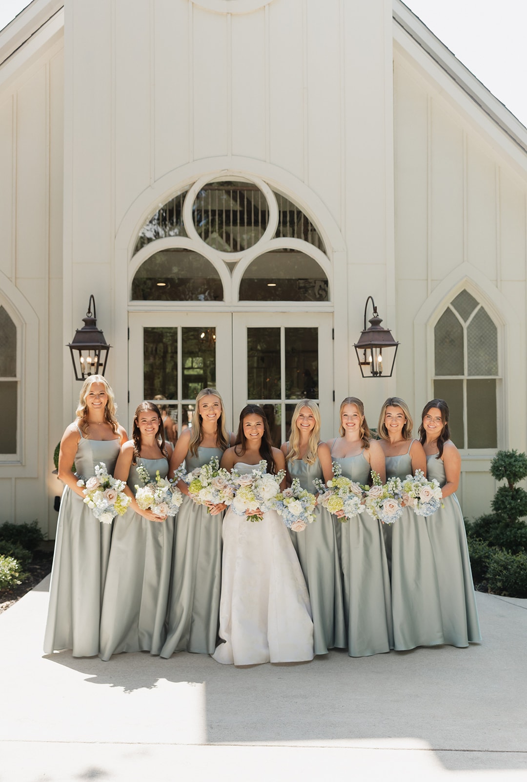 Nina and her seven bridesmaids in light blue dresses holding blush and white bouquets in front of the white French Farmhouse chapel exterior on July 4th, 2026