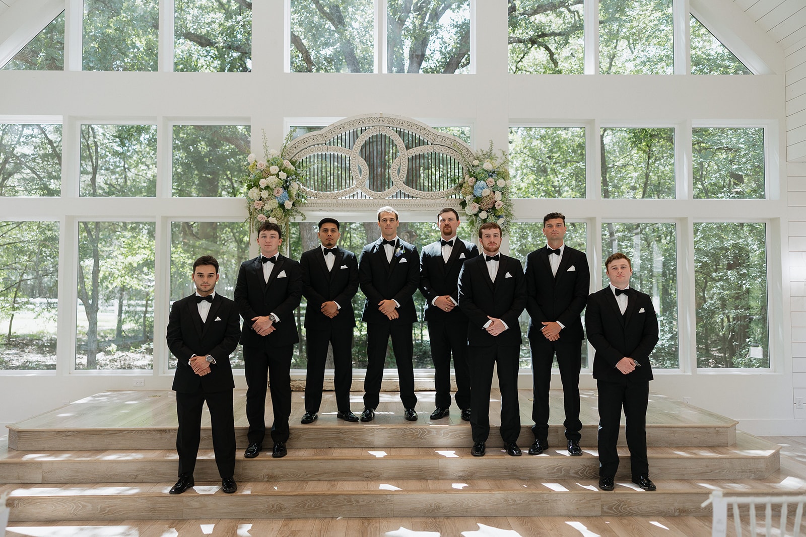 Gage and his seven groomsmen in classic black tuxedos lined up on the chapel altar steps at The French Farmhouse Venue with the antique arch and JG Floral blue and white arrangements behind them