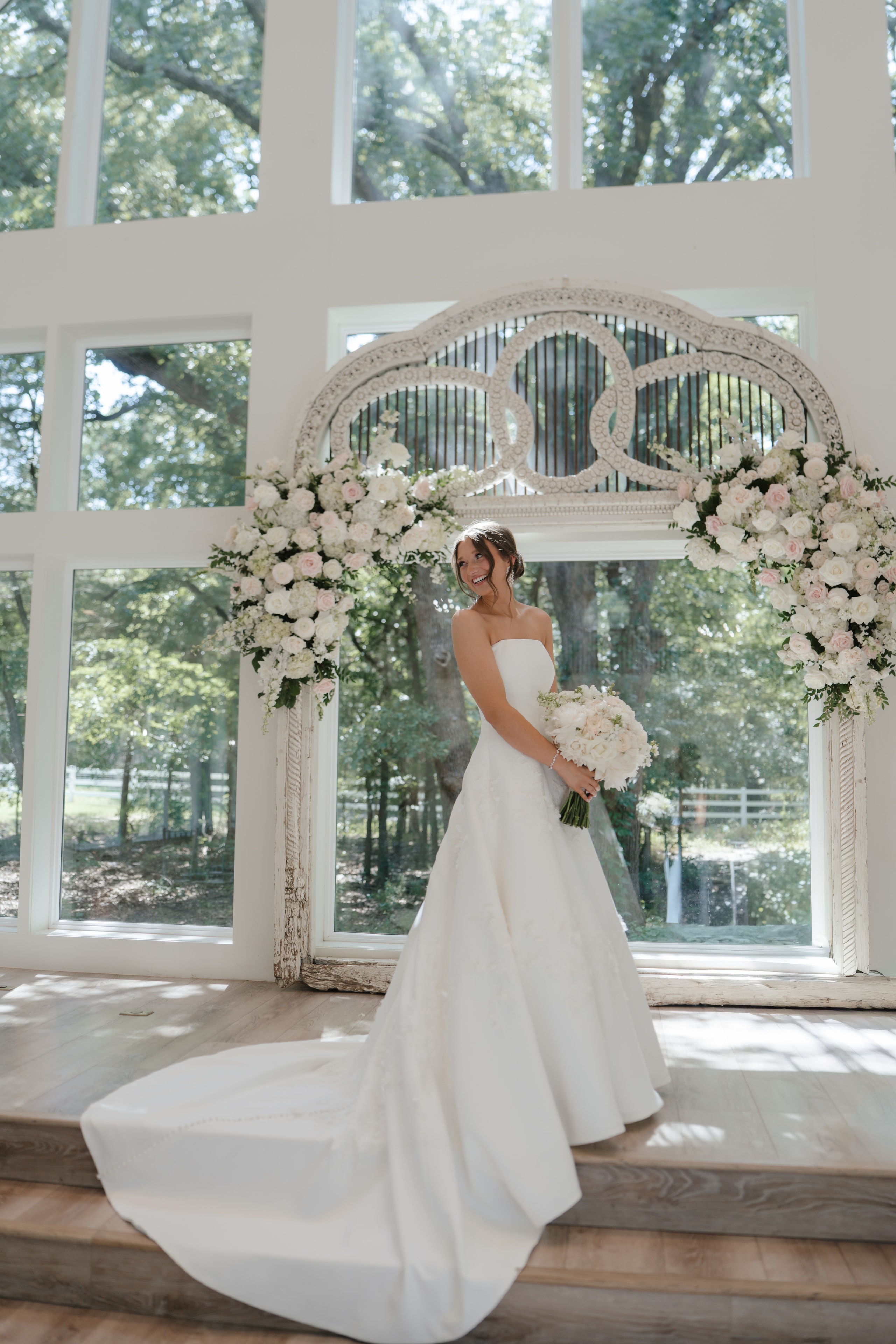 Paige in her strapless white gown standing in front of the antique arch adorned with white and blush roses inside The French Farmhouse Venue chapel