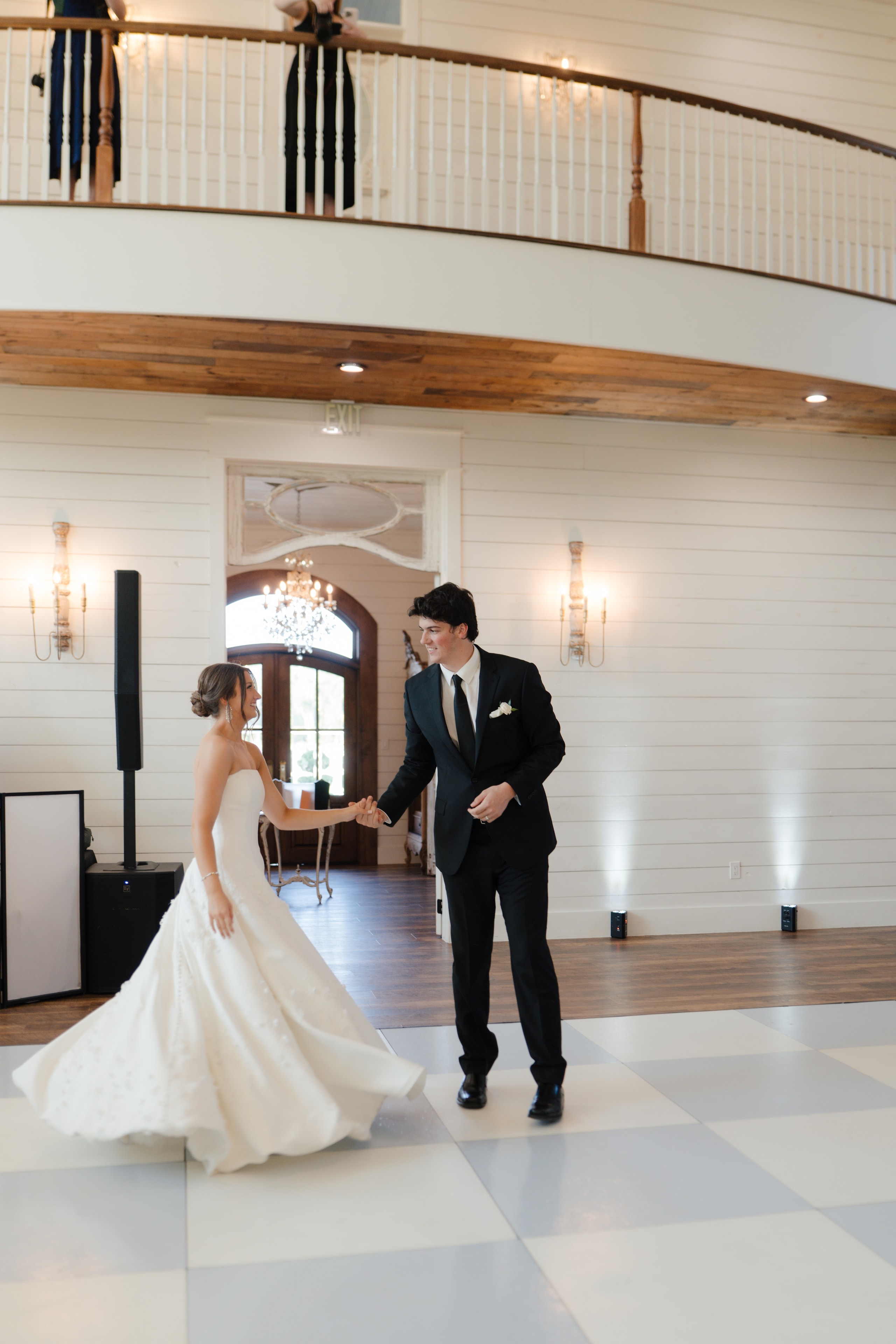 Paige and DJ sharing their first dance on the checkered dance floor beneath the balcony at The French Farmhouse Venue reception hall