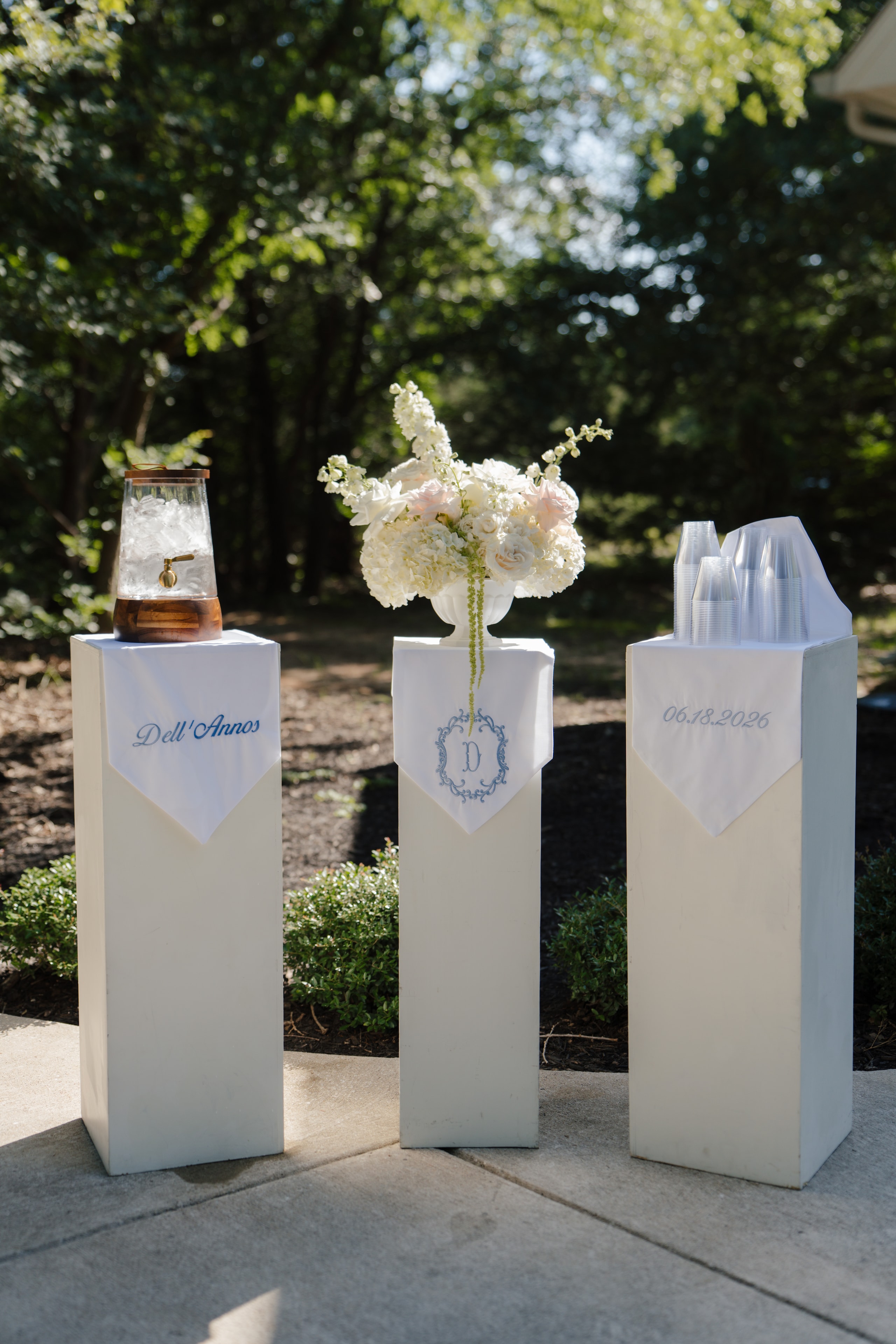 Elegant monogrammed water station with white pedestals, a floral arrangement, and embroidered linens reading Dell'Annos and 06.18.2026 outside the chapel at The French Farmhouse Venue