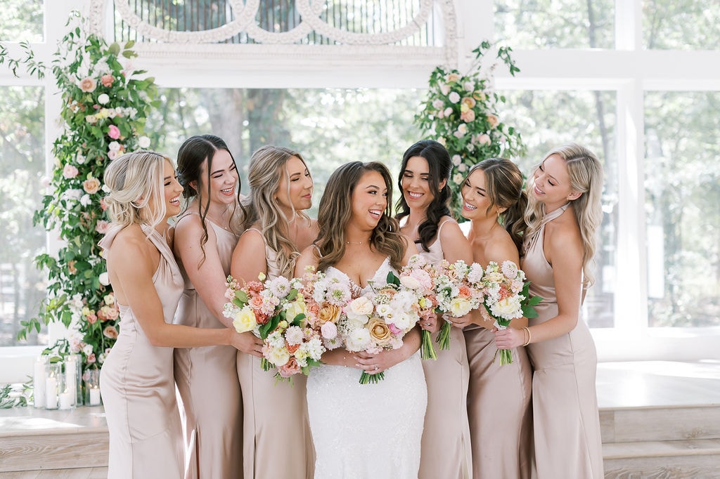 Shelby surrounded by her six bridesmaids in champagne satin gowns inside The French Farmhouse bridal suite, all laughing and looking at each other, holding lush bouquets of coral, blush, peach, and cream garden roses, dahlias, and ranunculus, the ornate white mirror and floor-to-ceiling windows with oak trees glowing behind them