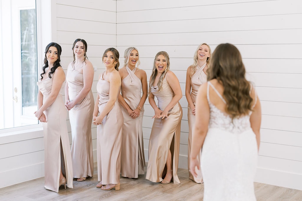 Six bridesmaids in champagne satin gowns lined up against the white shiplap wall of The French Farmhouse bridal suite, mouths open and screaming with joy as Shelby in her white lace wedding gown turns to reveal herself for the first time, the reaction completely unscripted and electric