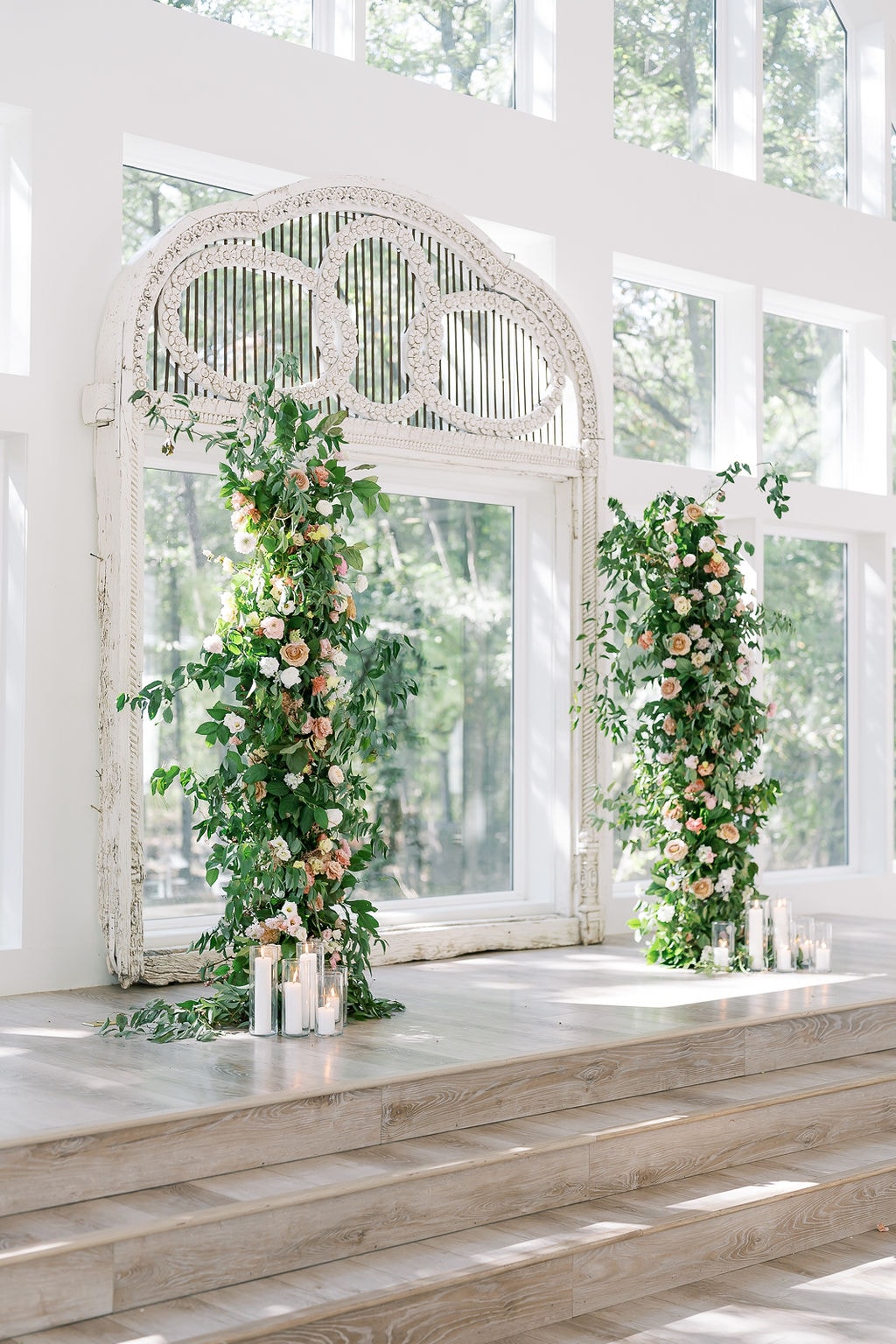 Close-up of the antique carved white arch at the altar of The French Farmhouse chapel, draped in lush climbing greenery, blush garden roses, peach ranunculus, and white flowers cascading down both sides, pillar candles in glass cylinders at the base, the floor-to-ceiling windows and summer oak trees glowing in the background