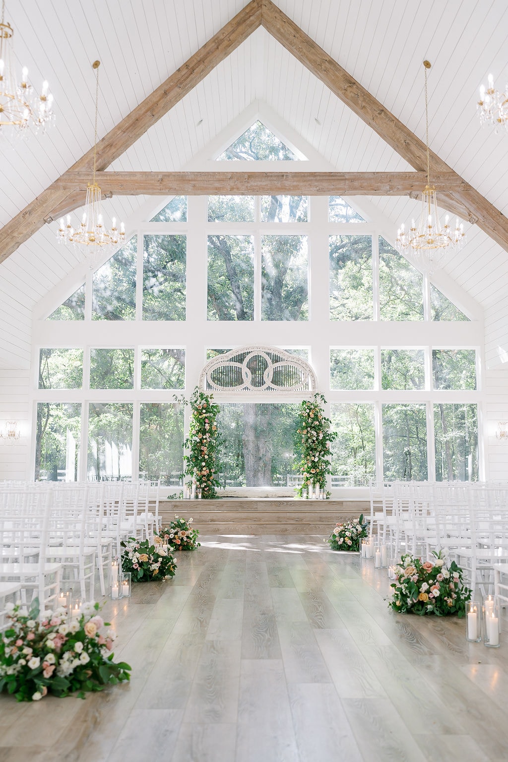 Wide shot of The French Farmhouse chapel set for Shelby and Cole's ceremony — soaring cathedral ceilings with exposed wood beams, crystal chandeliers, white Chiavari chairs lining the aisle, lush blush and peach floral clusters with pillar candles at each row end, the antique carved white arch flanked by tall floral columns at the altar, floor-to-ceiling windows with summer oak trees glowing behind