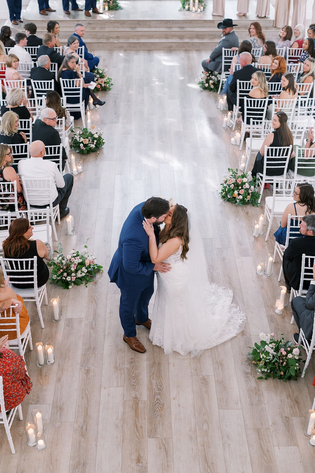 Aerial shot looking straight down the aisle of The French Farmhouse chapel as Shelby and Cole share their first kiss as husband and wife, blush and peach floral clusters and pillar candles lining both sides of the aisle, guests in black tie seated in white Chiavari chairs on either side, the light wood floors and soft natural light creating a dreamy overhead perspective
