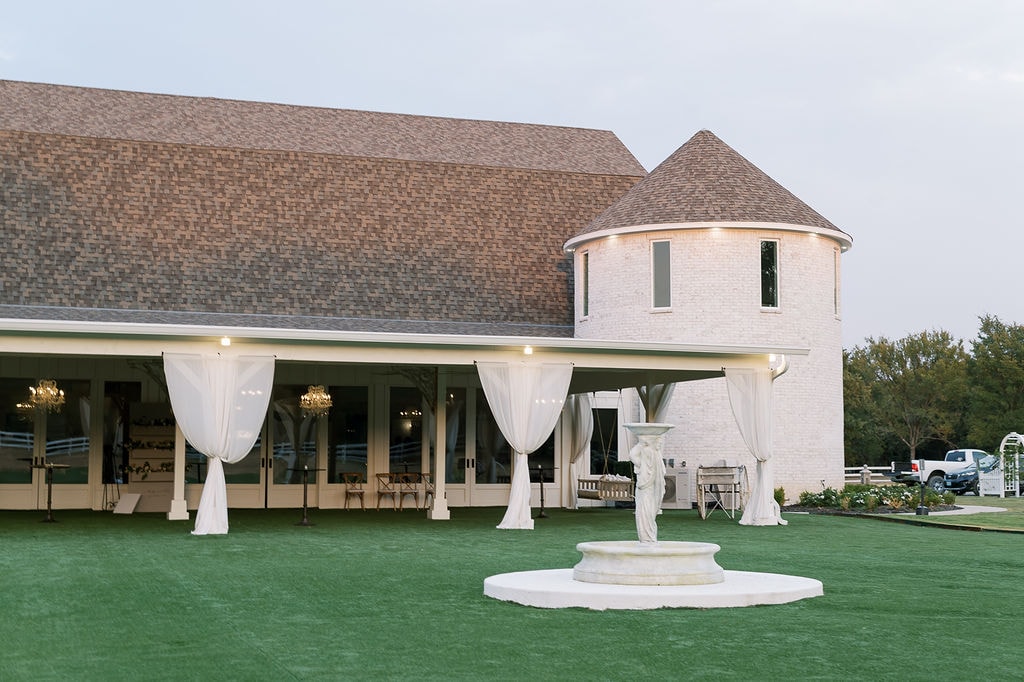 Dusk shot of The French Farmhouse Venue cocktail hour — the white brick pavilion with white linen draping tied back at each column, gold chandeliers glowing inside, the round turret tower lit from above, and the white stone angel fountain centered on the lush green lawn in the foreground, oak trees and white fence lining the property in the background