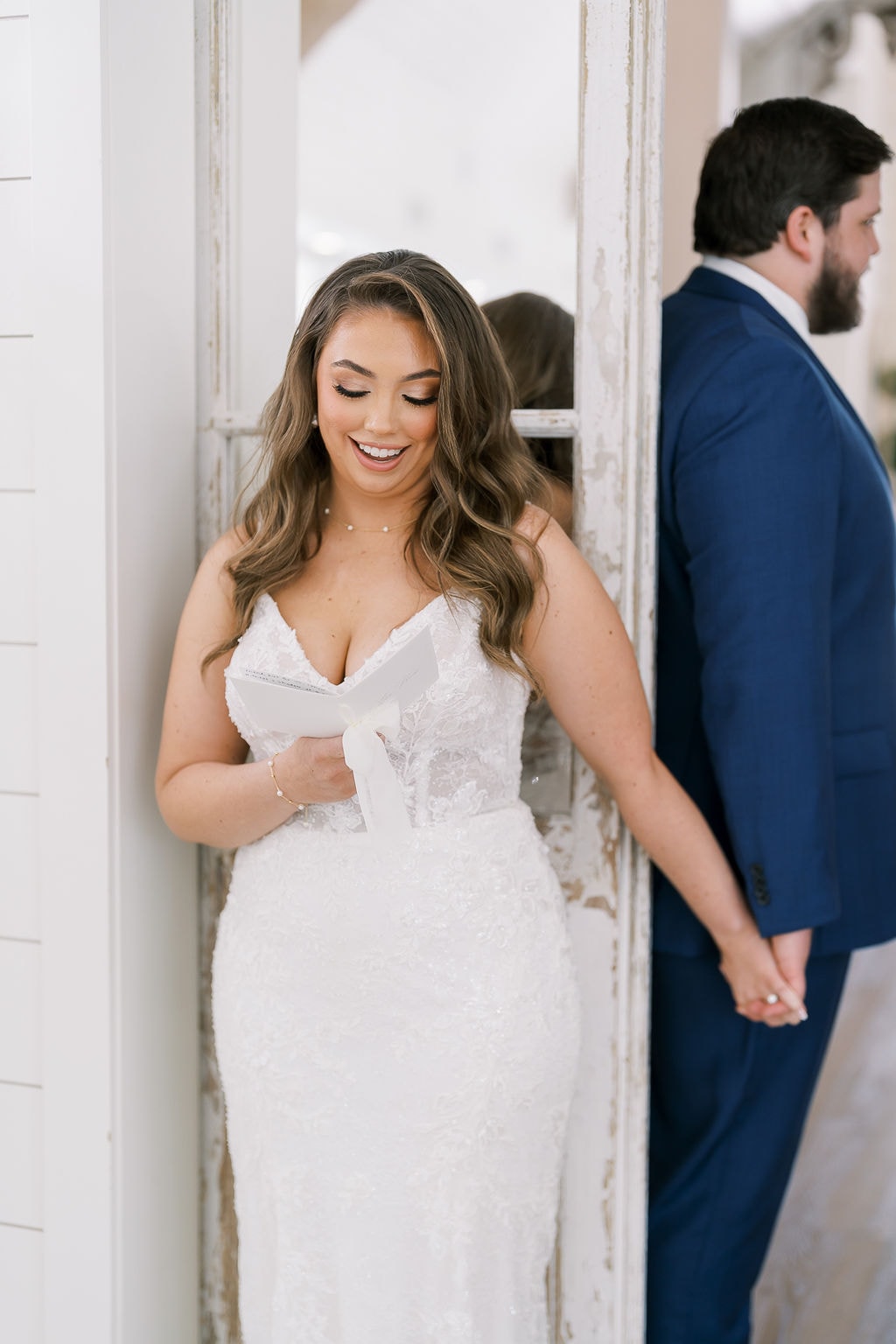 Close-up of Shelby beaming as she reads Cole's private vow letter, her lace fitted gown and pearl necklace glowing in the soft natural light, Cole in his navy suit visible behind her through the antique door frame, their hands still intertwined