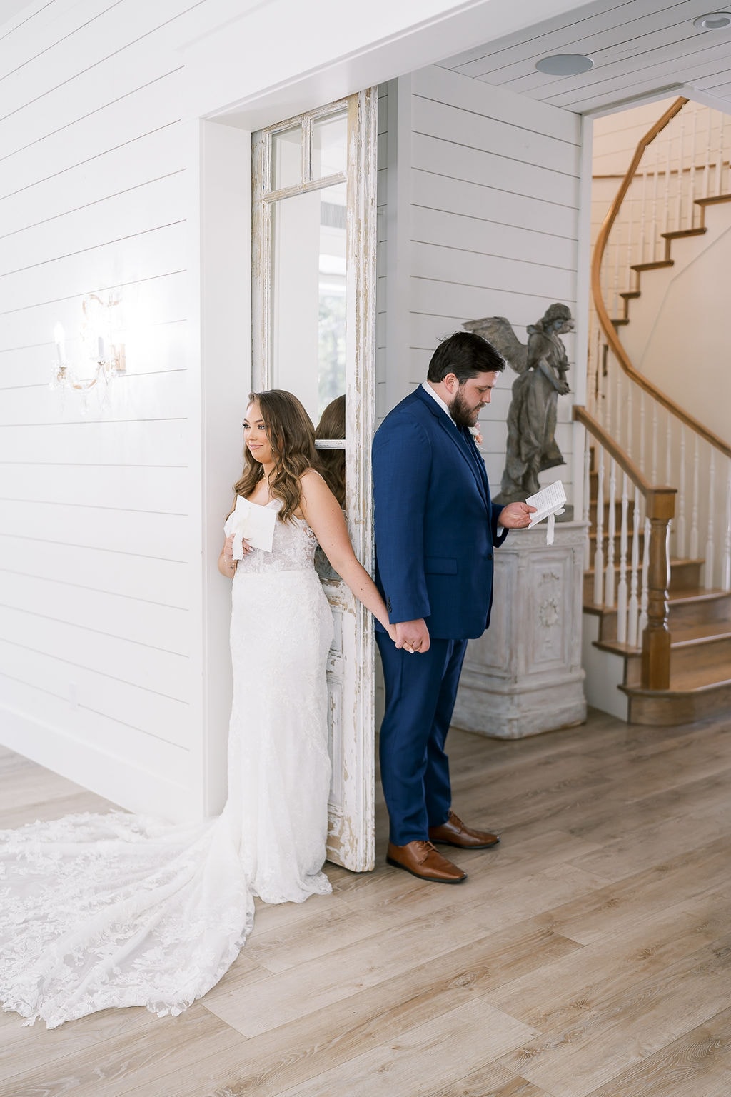 Shelby and Cole standing back to back on either side of the antique distressed white door inside The French Farmhouse, holding hands around the door frame while reading their private vow letters, the curved staircase and angel statue visible behind Cole, Shelby smiling softly at her letter in her lace fitted gown