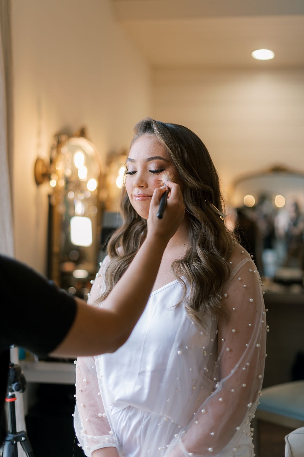 Shelby having her makeup applied inside The French Farmhouse bridal suite, wearing a white pearl-embellished robe, her long brunette waves styled down, the ornate gold vanity mirror and warm Edison bulb sconces glowing behind her