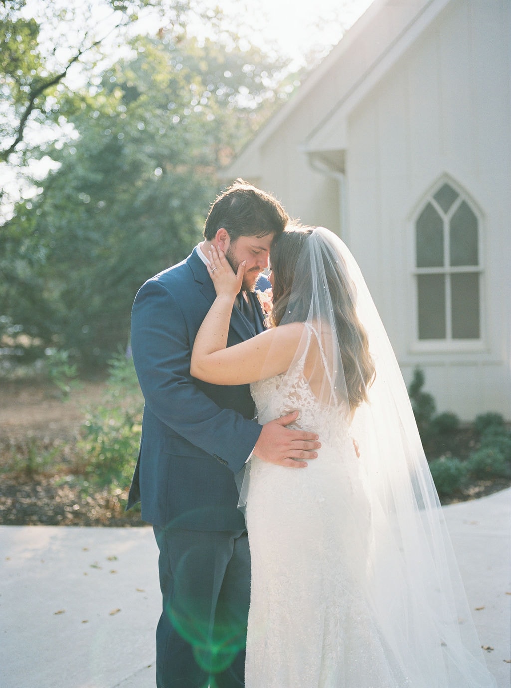 Shelby and Cole embracing in front of The French Farmhouse chapel exterior, Cole holding her close with his hand on her back, Shelby's cathedral veil billowing behind her lace gown, the white board-and-batten chapel with Gothic arched windows and mature oak trees glowing in warm golden afternoon light behind them