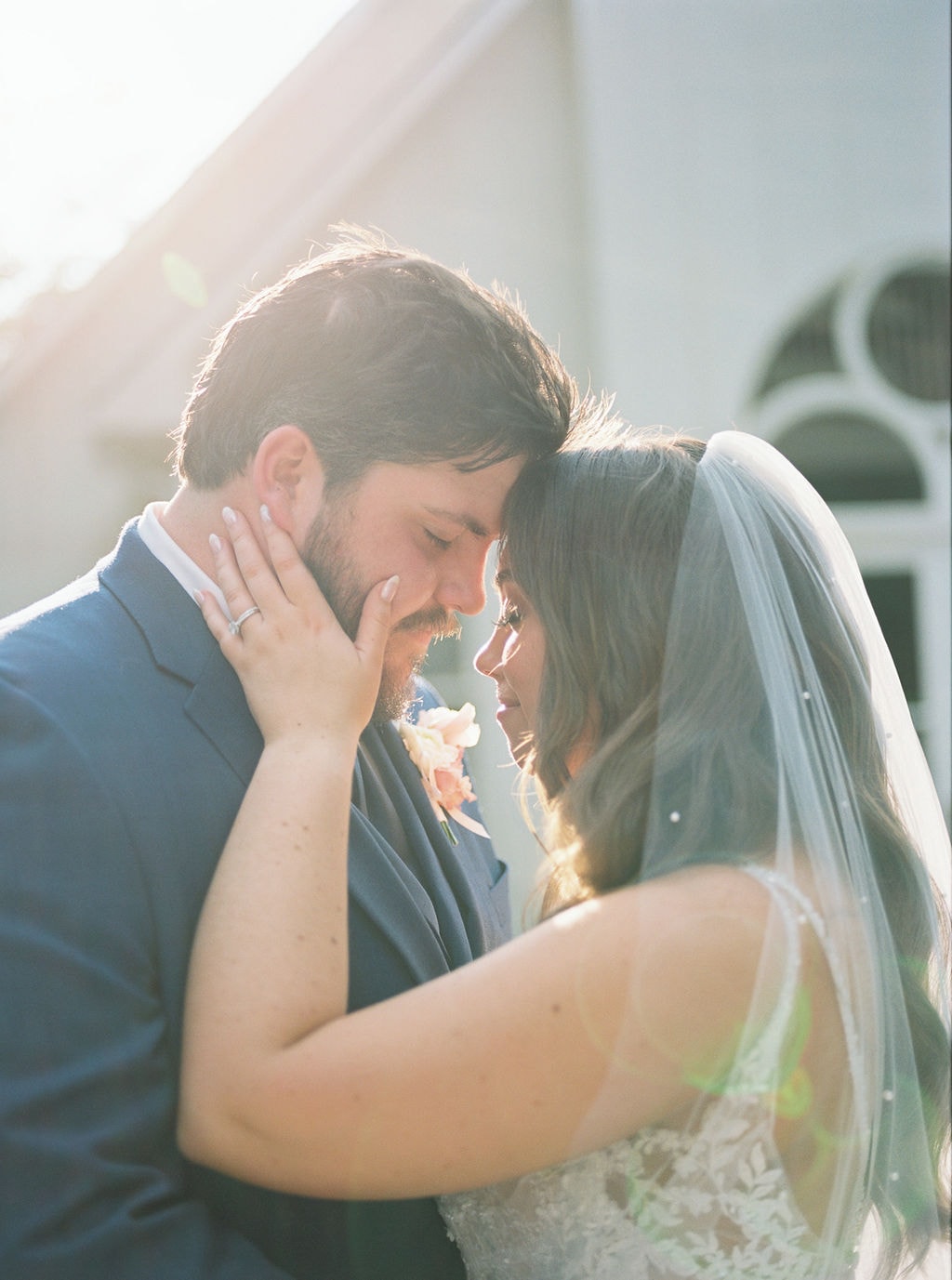 Close-up of Shelby and Cole pressing their foreheads together outside The French Farmhouse chapel, Shelby's hand cradling Cole's face, her pearl-edged veil cascading over her lace gown, Cole's navy suit boutonniere with a blush peach rose, warm golden backlight flooding the frame with a soft lens flare