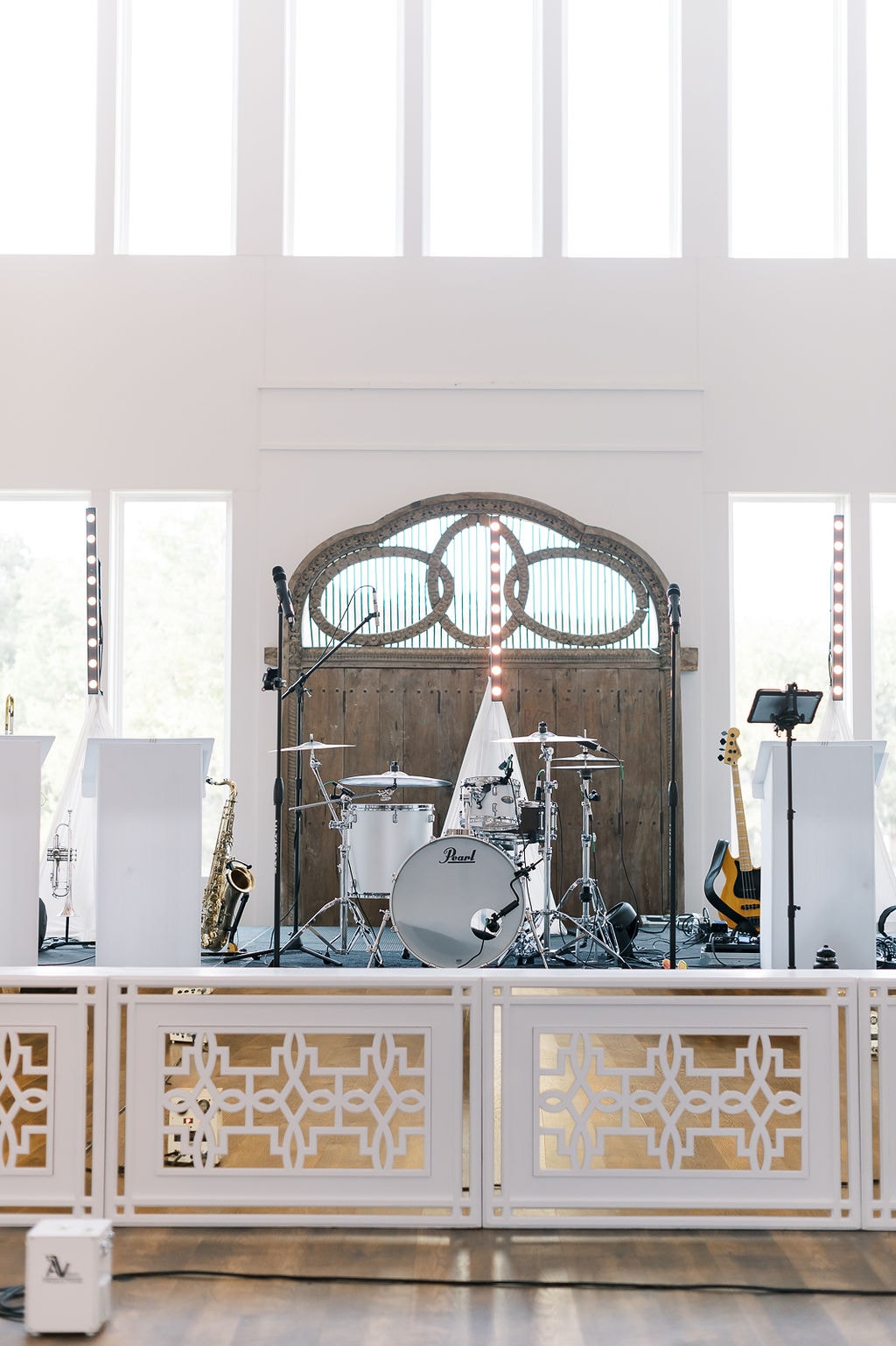 The Jordan Kahn Orchestra stage set up inside The French Farmhouse reception hall — Pearl drum kit, saxophone, trumpet, and guitars arranged in front of the ornate antique carved wood barn doors with marquee bulb lighting, white and gold geometric stage facade panels in the foreground, soaring floor-to-ceiling windows flooding the space with natural light