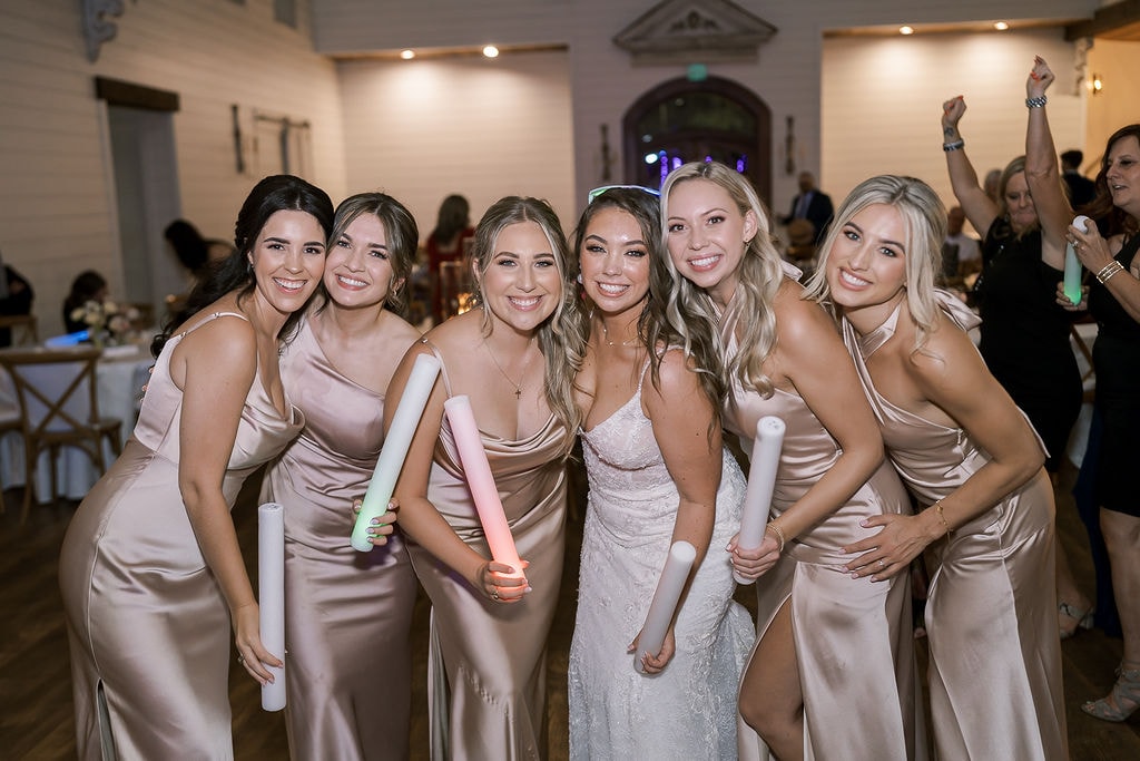 Shelby beaming in the center of her bridesmaids in champagne satin gowns on the dance floor inside The French Farmhouse reception hall, all holding glowing foam batons and glow sticks, laughing and posing for the camera, guests dancing in the background under the warm Edison bulb lighting