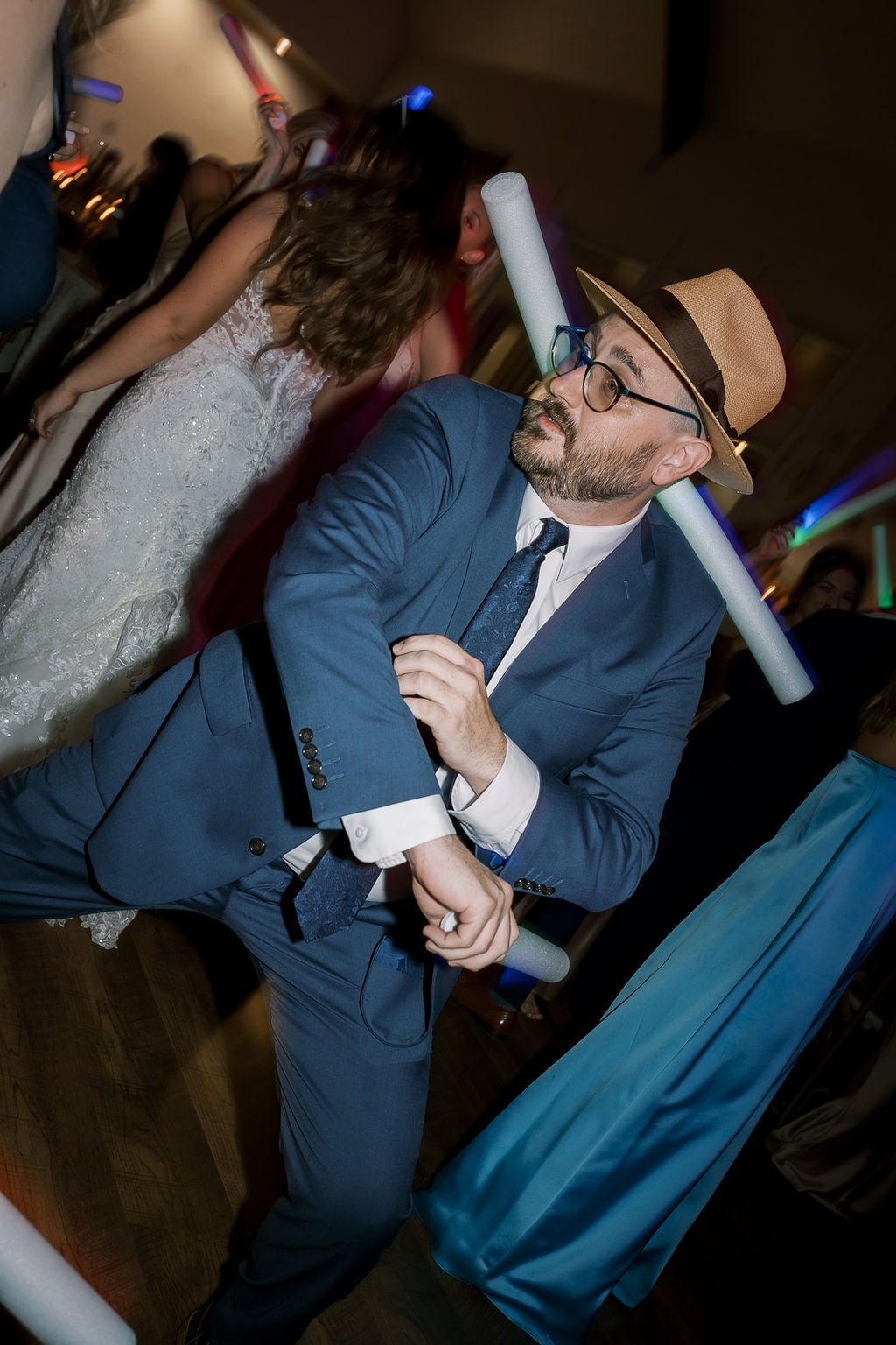 A guest in a navy suit and straw fedora completely losing himself on the dance floor at The French Farmhouse reception, mid-spin with a foam baton balanced on his shoulder, colorful uplighting washing the room in blue and purple, Shelby in her lace gown visible dancing beside him