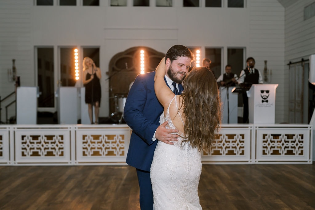 Shelby and Cole sharing an intimate last dance together on the empty dance floor inside The French Farmhouse reception hall, the Jordan Kahn Orchestra glowing on stage behind them in front of the antique barn doors, warm Edison bulb lighting washing the room in gold