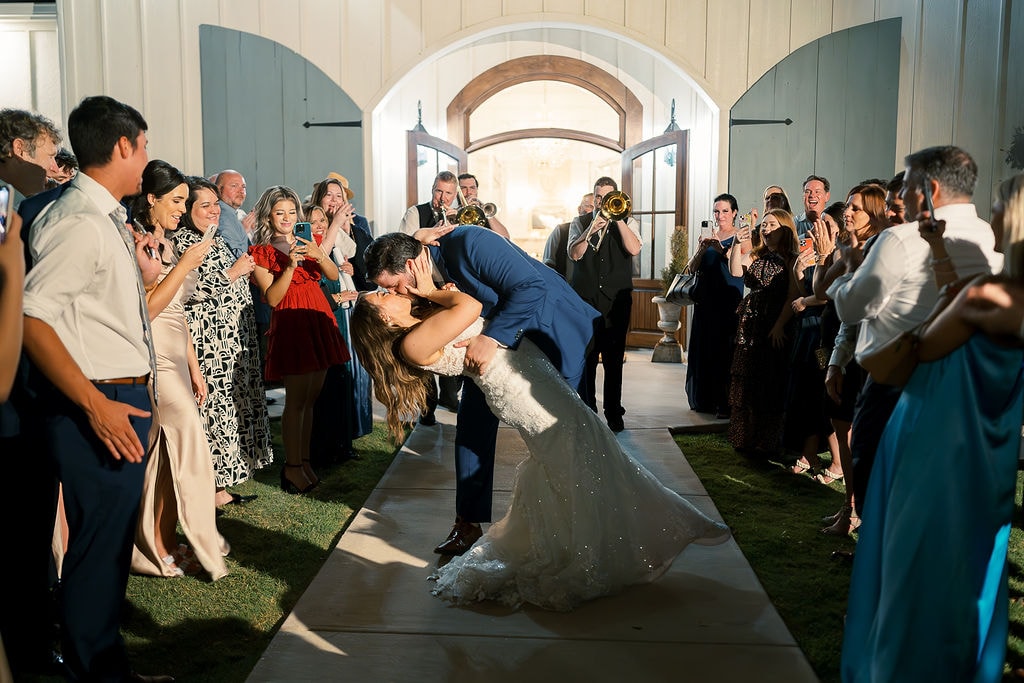 Cole dipping Shelby into a dramatic kiss on the courtyard pathway outside The French Farmhouse arched entry doors during their send-off, the Jordan Kahn trumpet player in a vest leading them out, the entire crowd of guests lining both sides cheering and filming on their phones, the sage blue shutters and white board-and-batten facade glowing behind them at night