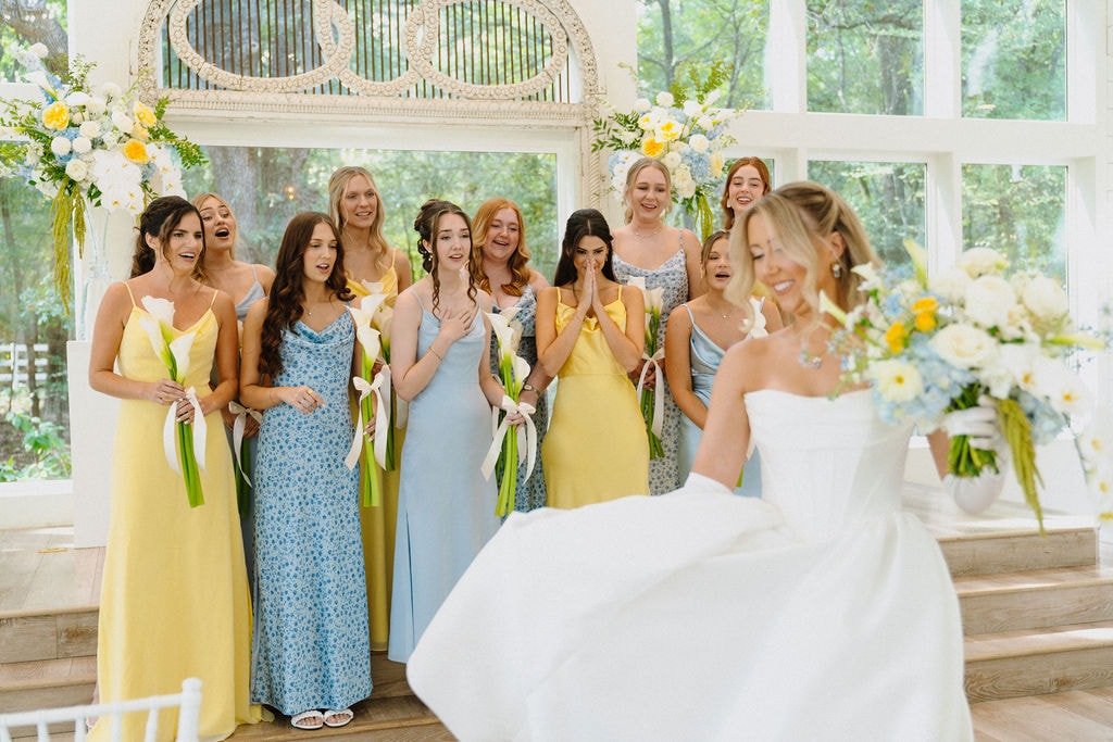 Bride revealing her dress to bridesmaids in yellow and blue gowns inside the chapel at The French Farmhouse Venue