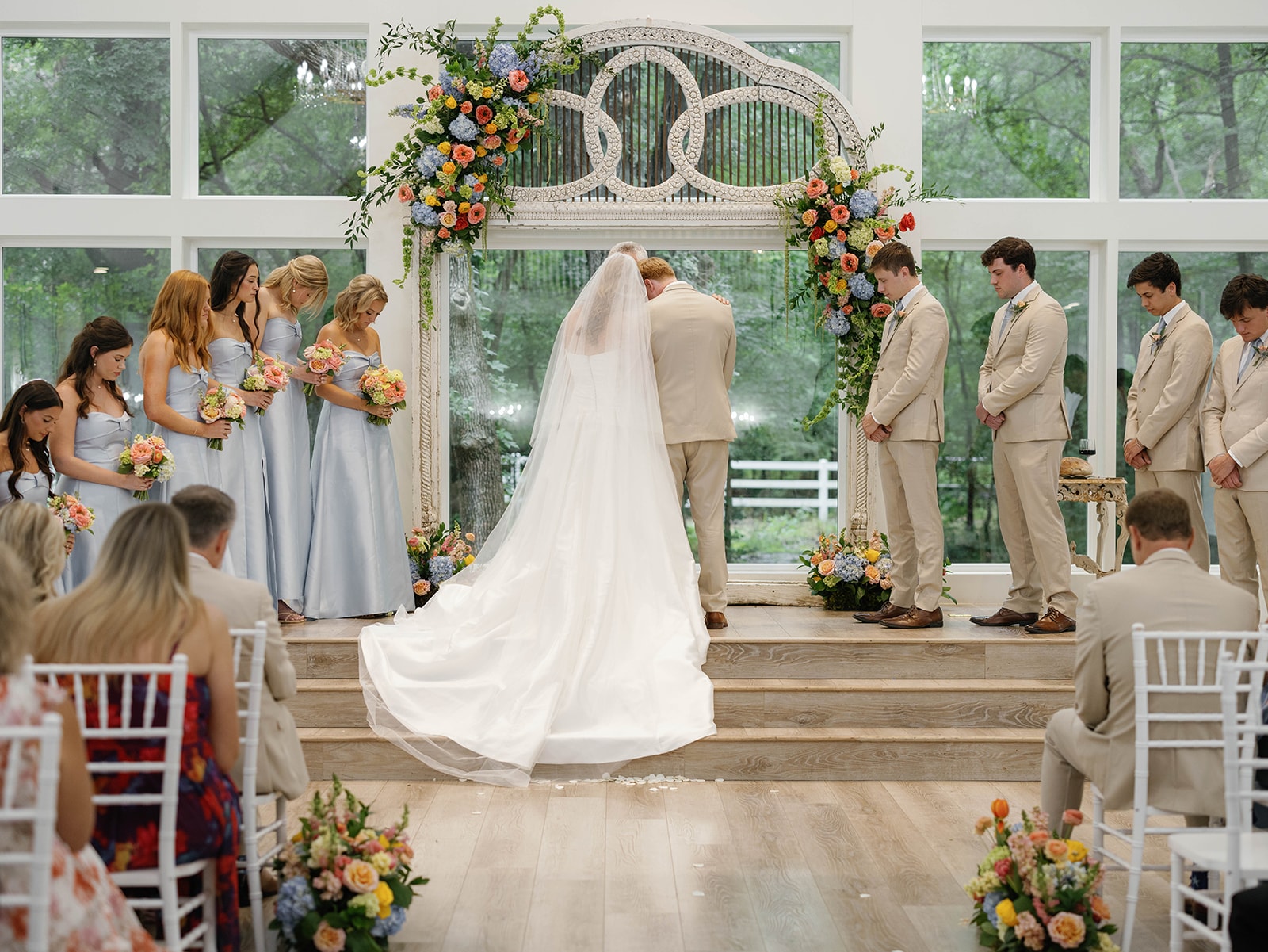 Bride and groom praying together at the altar with bridal party and colorful floral arch at The French Farmhouse Venue