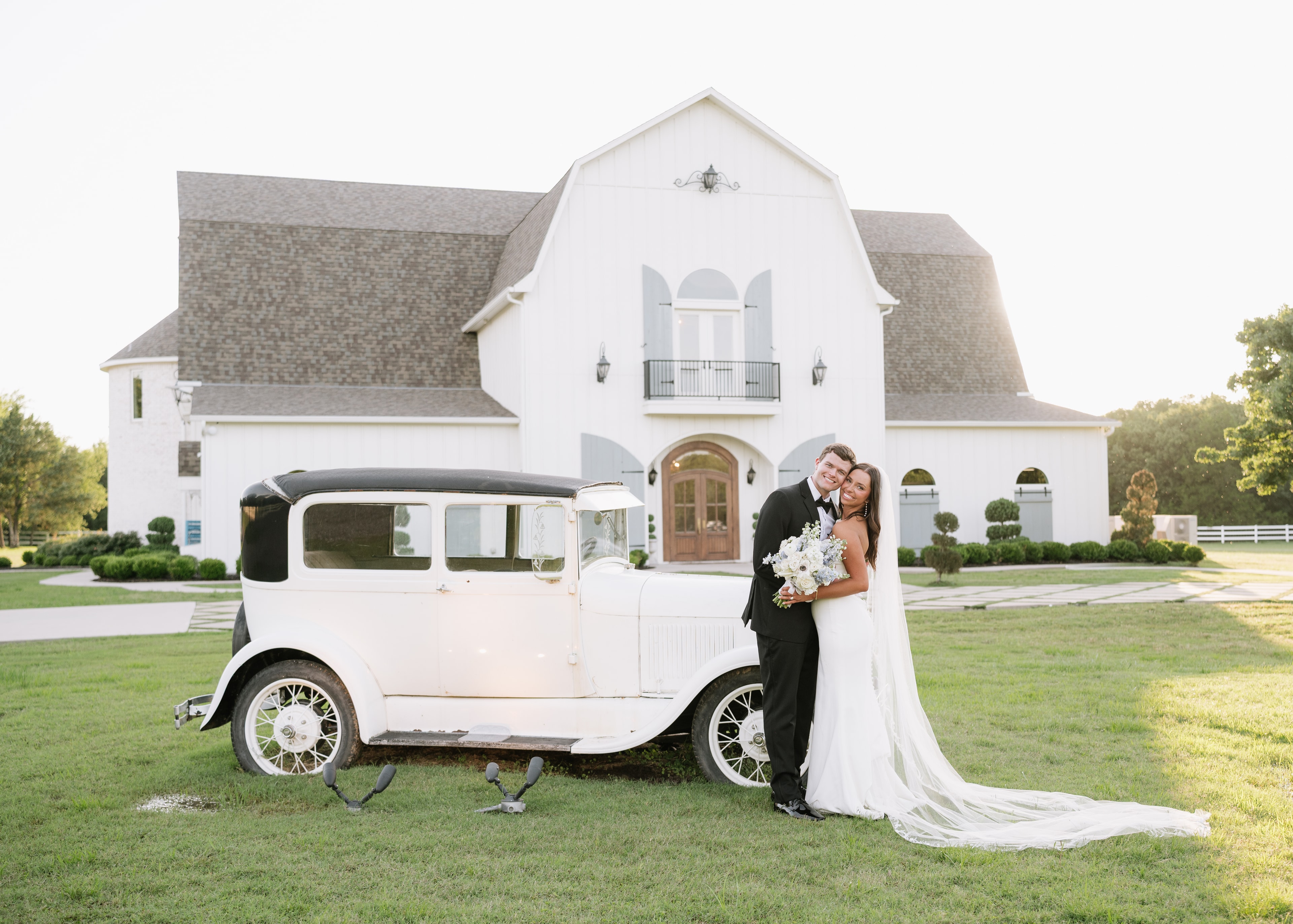Bride and groom posing with a vintage white car in front of The French Farmhouse Venue at golden hour