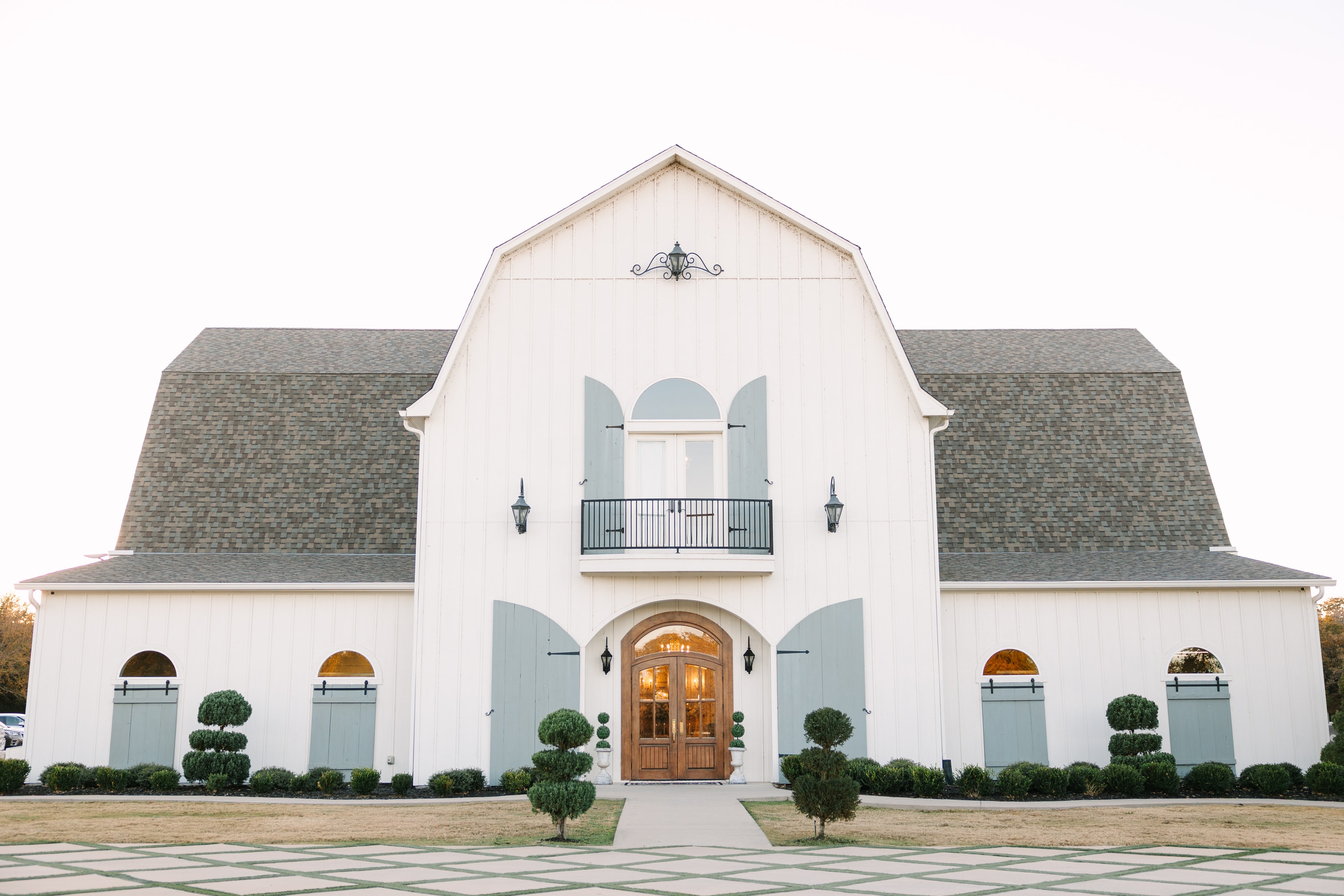 The French Farmhouse Venue exterior — white barn facade with sage shutters, arched wood doors, topiary shrubs, and a glowing interior at golden hour in Collinsville, Texas
