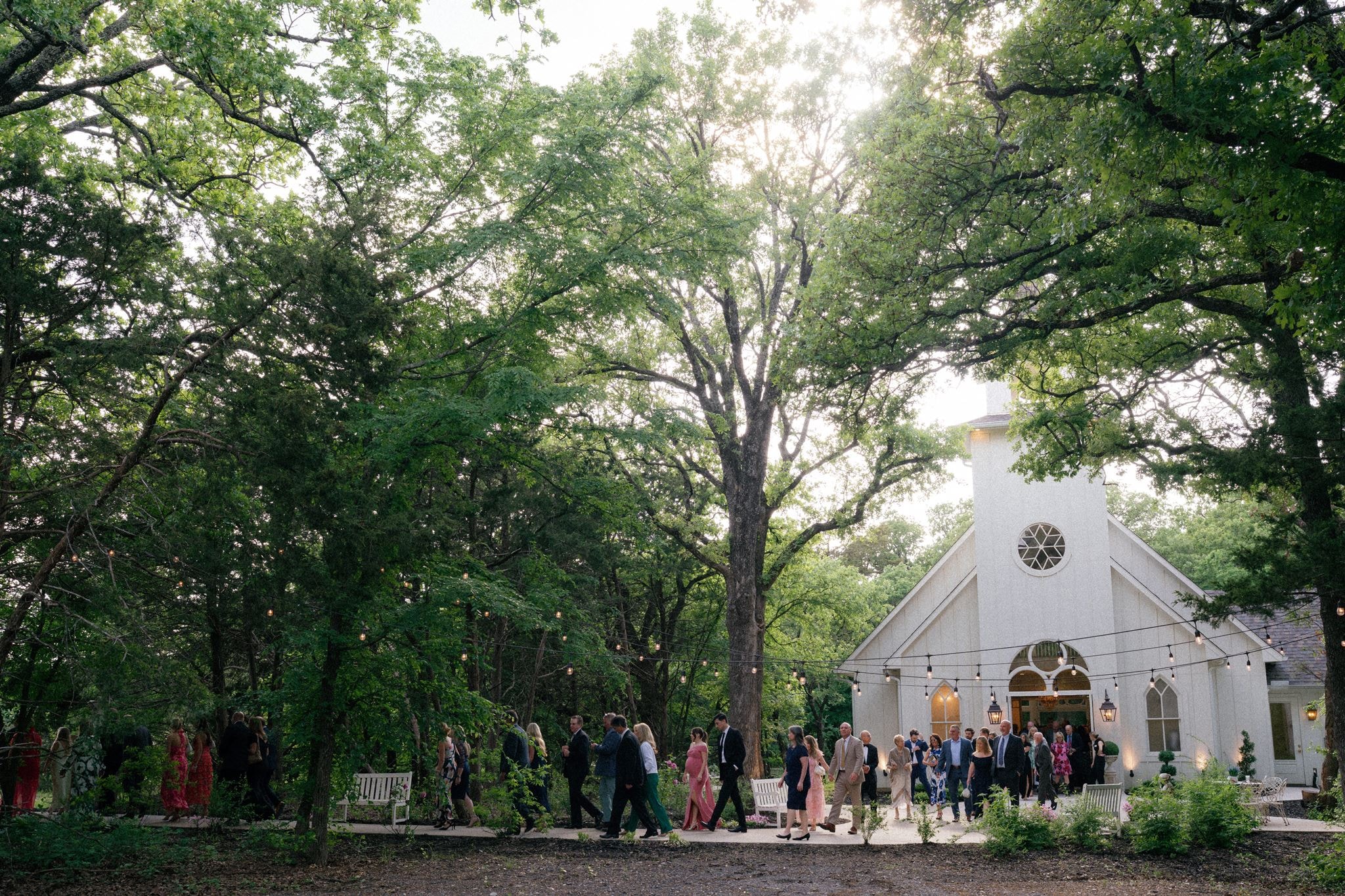 Wedding guests gathering outside The French Farmhouse chapel surrounded by trees and string lights at dusk