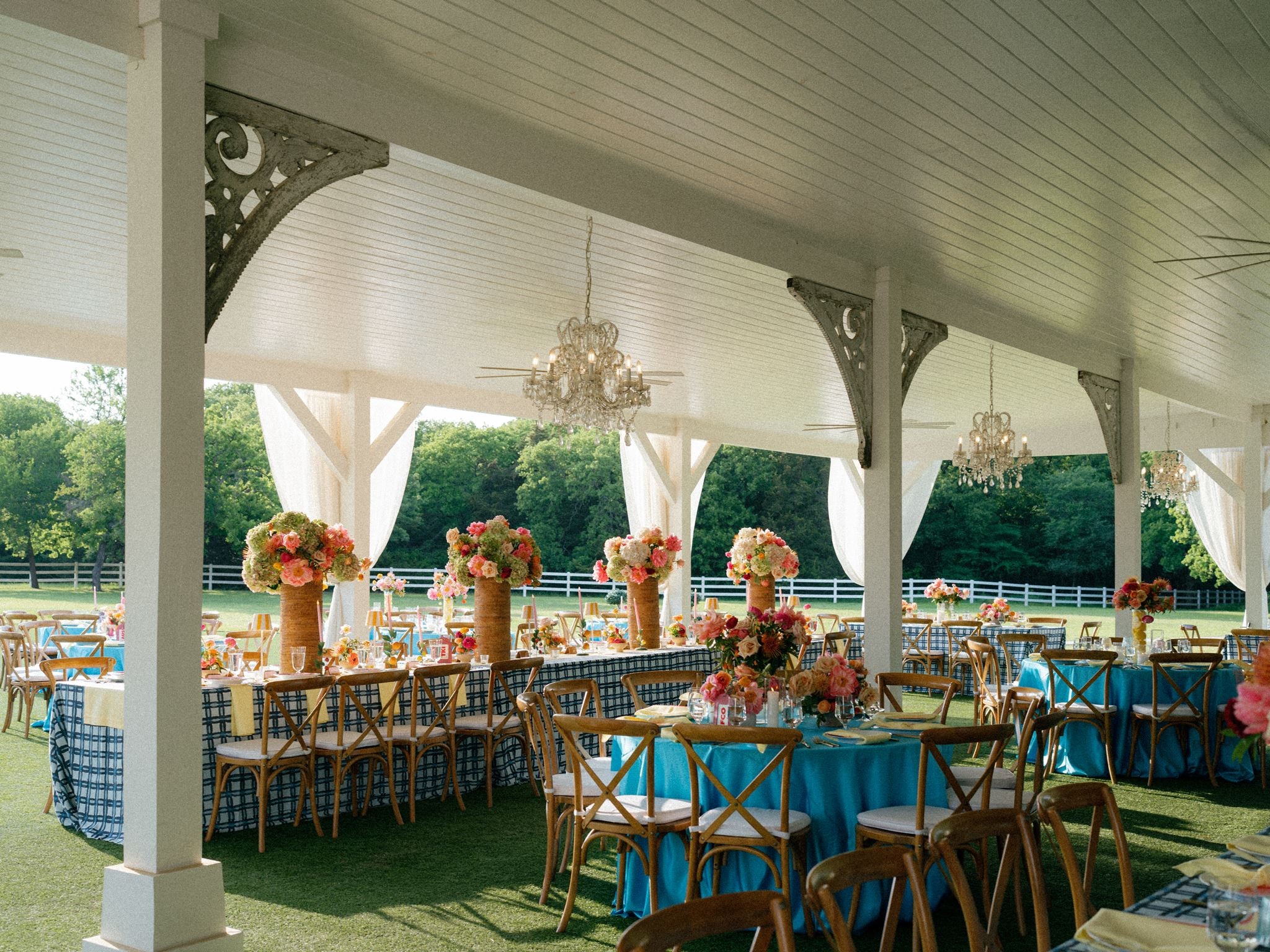 Colorful outdoor pavilion reception setup with blue linens, floral centerpieces, and chandeliers at The French Farmhouse Venue